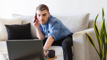 Retrato de un elegante y apuesto joven adulto caucásico trabajando desde su oficina en su hogar en una brillante y cálida habitación ocupado mientras usa su laptop y maneja su telefono movil