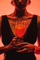 Wall Mural - cropped view of elegant woman holding glass with mixed cocktail and orange on pink