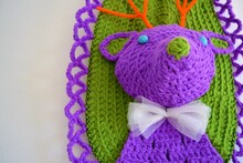 Closeup Of A Crocheted Taxidermy Deer In Purple And Green With A White Bow Tie Hanging On A Wall