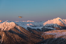 A Single Engine Airplane Flying Through Snowy Mountains At Sunset