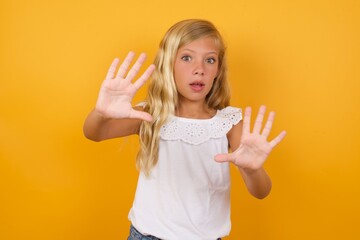 Wall Mural - Afraid Beautiful Caucasian young girl standing against yellow background, makes terrified expression and stop gesture with both hands saying: Stay there. Panic concept.