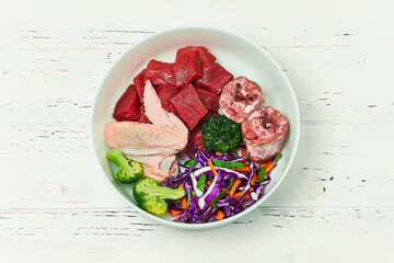 Wall Mural - Pet food. Bowl of natural raw dog food on white wooden background. Beef meat, turkey neck, chicken wing and vegetables. BARF diet for dog.