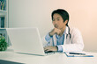 © steafpong - Serious Young Asian Doctor Man in Lab Coat or Gown with Stethoscope Touch Chin and Using Laptop Computer on Doctor Table in Office in Vintage Tone