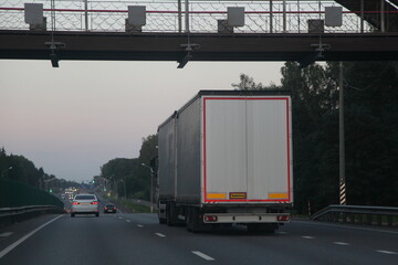 European white van truck with semi trailer drive on two lane suburban asphalted highway under metal bridge, back view at summer evening , road transportation cargo logistics