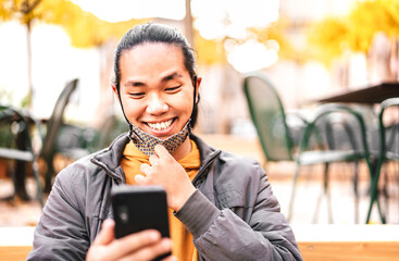 Happy asian guy smiling with open face mask after lockdown reopening - New normal lifestyle concept by confident young man watching news on mobile smart phone - Warm bright filter with focus on eyes