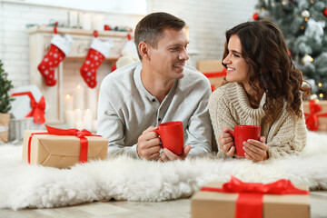 Canvas Print - Happy couple with cups of hot drink on floor in room decorated for Christmas