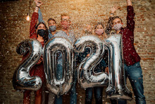 Happy Group Of Friends With Face Mask Celebrating New 2021 Year - Confetti Falling In The Air - Young People With Balloons In Hands Making New Year Party At Home - Focus On Balloons.