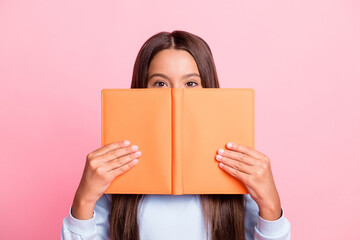 close-up portrait of pretty funny brown-haired girl reading hiding behind book isolated over pink pa