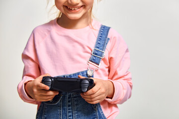 Cute kid girl playing video games holding gamepad and smiling , wearing pink casual sweatshirt and jeans jumpsuit, on white background