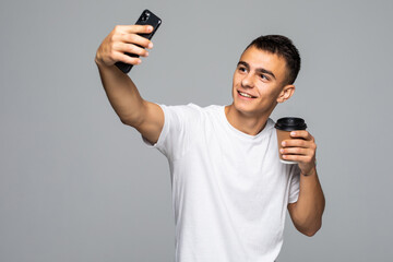 Wall Mural - Handsome confident young man standing isolated over gray background, taking a selfie, holding cup of takeaway coffee