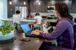 © Inti St. Clair - Woman working from home on laptop computer with puppy in lap while daughters study at kitchen counter in background