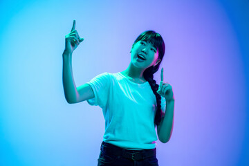 Wall Mural - Pointing up. Young asian woman's portrait on gradient blue-purple studio background in neon light. Concept of youth, human emotions, facial expression, sales, ad. Beautiful brunette model.