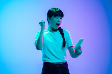 Wall Mural - Winner with phone. Young asian woman's portrait on gradient blue-purple studio background in neon light. Concept of youth, human emotions, facial expression, sales, ad. Beautiful brunette model.