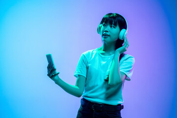 Wall Mural - Listening to music, dancing in headphones. Young asian woman's portrait on gradient blue-purple studio background in neon. Concept of human emotions, facial expression, sales, ad. Beautiful model.