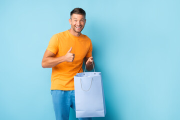 cheerful man in t-shirt holding shopping bag and showing thumb up on blue