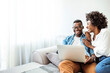 Young black couple using laptop sitting on the sofa at home. Full length of couple watching movie on laptop. Happy man and woman are relaxing on couch. They are spending leisure time at home.