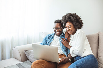 Young black couple sit on the sofa using laptop. Black couple using laptop at home look at each other. Overjoyed african American couple sit relax on cozy couch look at laptop screen triumph winning 