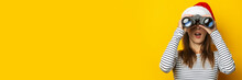 Young Woman With A Surprised Face In A Santa Claus Hat Looks Through Binoculars On A Yellow Background. Banner