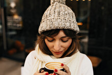 Outside Portrait Of Pretty Charming Girl With Coffee Wearing In Winter Cap And White Sweater In Lights. Christmas Mood
