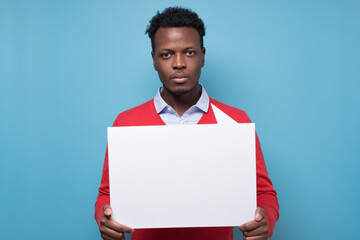 Wall Mural - Handsome serious african business man presenting some product on empty placard. Studio shot on blue wall.