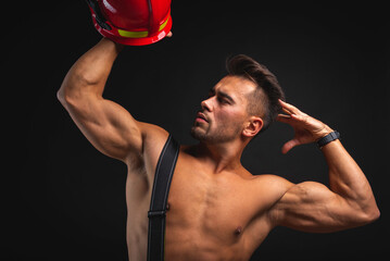 Wall Mural - young handsome adult firefighter in uniform and helmet holding firefighting equipment helmet in hands, isolated on dark background 