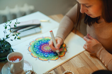 Young woman colouring mandala with markers and white rosary on table with cup of coffee at home