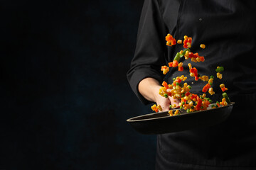 Wall Mural - Chef throws up frying mix of colored vegetables above the pan on dark blue background. Backstage of cooking meal. Frozen motion. Food banner concept.