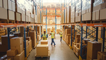 Retail Warehouse Full Of Shelves With Goods In Cardboard Boxes, Workers Scan And Sort Packages, Move Inventory With Pallet Trucks And Forklifts. Product Distribution Logistics Center. Elevated Shot