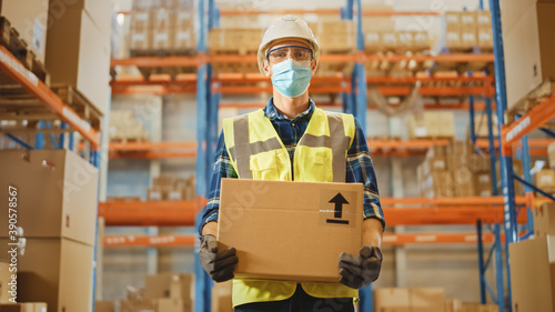 Handsome Male Worker Wearing Medical Face Mask and Hard Hat Carries Cardboard Box Stands in Retail Warehouse full of Shelves with Goods. Logistics, Distribution Center. Fromt Shot