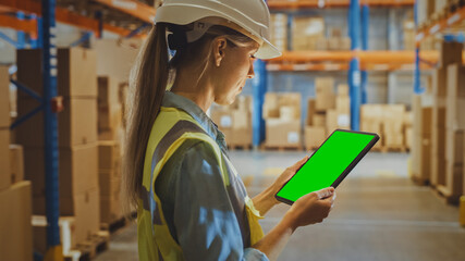 Wall Mural - Professional Female Worker Wearing Hard Hat Uses Digital Tablet Computer with Green Chroma Key Screen in Portrait Mode in the Retail Warehouse full of Shelves with Goods. Logistics and Distribution 