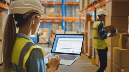 Professional Female Worker Wearing Hard Hat Holds Laptop Computer with Screen Showing Inventory Checking Software in the Retail Warehouse full of Shelves with Goods. Over the Shoulder Side View 