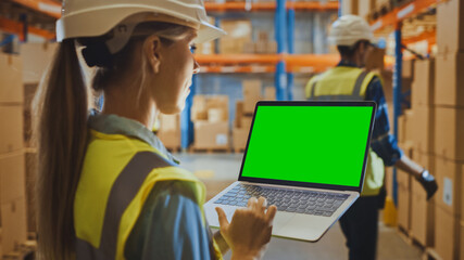 Wall Mural - Professional Female Worker Wearing Hard Hat Holding Laptop Computer with Green Chroma Key Screen in Landscape Mode in the Retail Warehouse full of Shelves with Goods. Over the Shoulder Side view 