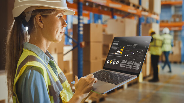 Professional Female Worker Wearing Hard Hat Holds Laptop Computer with Screen Showing Analysis Software in the Retail Warehouse full of Shelves with Goods. Over the Shoulder Side View 