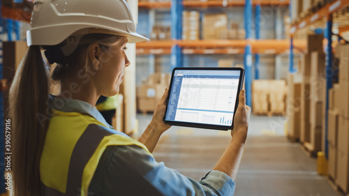 Professional Female Worker Wearing Hard Hat Uses Digital Tablet Computer with Screen Showing Inventory Checking Software in the Retail Warehouse full of Shelves with Goods. Over the Shoulder View