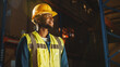 © Gorodenkoff - Handsome Professional Worker Wearing Safety Vest and Hard Hat Charmingly Smiling and Looks Into the Distance. In the Background Big Warehouse with Shelves full of Delivery Goods. Medium Portrait