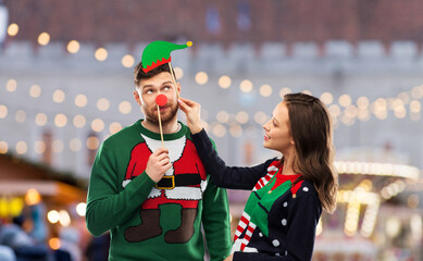 Wall Mural - people and holidays concept - happy couple in ugly sweaters posing with party props over christmas market background