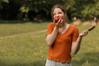 © Volodymyr - Close up lifestyle portrait of young girl college student devouring juicy red apple and holding ice lolly cream in hand. Woman in city park in sunny day. Image with blur background, copy empty space.
