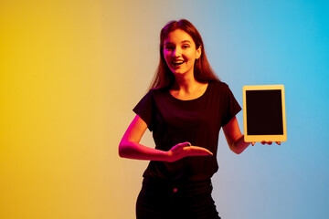 Wall Mural - Showing blank tablet screen. Young caucasian woman's portrait on gradient blue-yellow studio background in neon light. Concept of youth, human emotions, facial expression, sales, ad. Beautiful model.