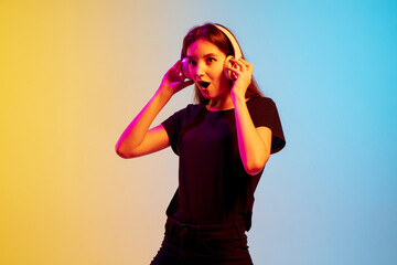 Wall Mural - Listening to music with headphones. Young caucasian woman's portrait on gradient blue-yellow studio background in neon light. Concept of youth, human emotions, facial expression, sales, ad.