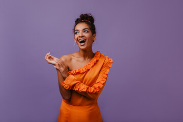 Cute african girl in orange dress posing with pretty smile. Debonair black female model laughing on purple background.