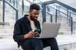 © F8  \ Suport Ukraine - Young African American man working on laptop computer, use phone, sitting on stairs
