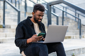 Young African American man working on laptop computer, use phone, sitting on stairs