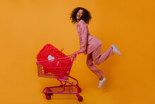 Enthusiastic Shopaholic Girl Jumping On Yellow Background. Trendy African Woman Posing With Shopping Cart.