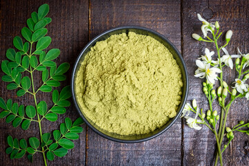 Wall Mural - Moringa powder in black bowl ceramic, fresh green leaves and Moringa flowers on wooden background.