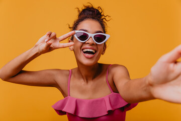 Spectacular young woman in sunglasses making selfie on yellow background. Indoor shot of laughing appealing african girl.