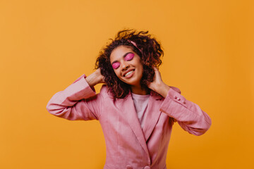 Wall Mural - Good-humoured wonderful woman in pink jacket enjoying photoshoot. Debonair young lady with curly hair dancing on orange background.