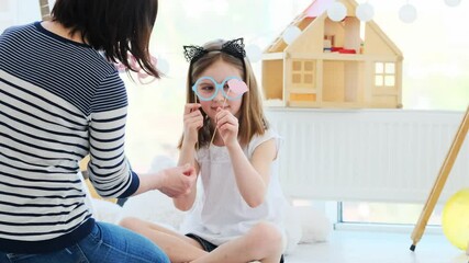 Wall Mural - mother and cute little daughter playing with paper glasses and lips sitting in kids room