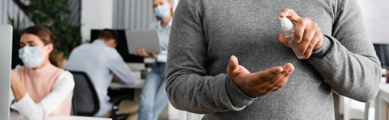 Cropped view of businessman using hand sanitizer near colleagues in medical masks on blurred background, banner