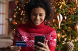 © fizkes - Close up smiling African American woman paying online by credit card for Christmas gifts, using smartphone, looking at screen, sitting near festive tree, shopping online, enjoying winter holiday
