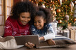 © fizkes - Close up happy African American woman with daughter using tablet together, lying on soft pillows near Christmas tree at home, happy family shopping online, choosing gifts, enjoying winter holiday